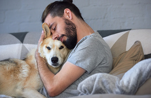 Ein dunkelhaariger, bärtiger Mann kuschelt mit einem großen Hund, der in die Kamera blickt.